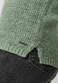 Maglione a maglia verde menta con un motivo testurizzato e spacchi laterali. Caratterizzato da un piccolo etichetta di marca sull'orlo, abbinato a jeans scuri.