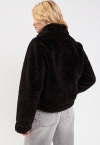 Schwarze Teddyjacke mit weichem, strukturiertem Finish. Verfügt über einen Kragen und eine lockere Passform, kombiniert mit hellgrauen Jeans im lässigen Stil.