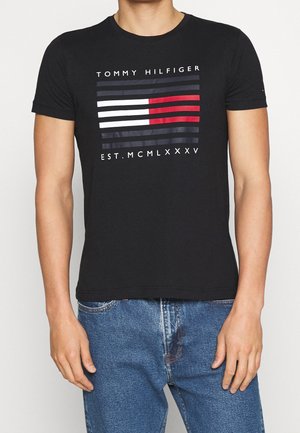 Mand iført sort Tommy Hilfiger T-shirt med vandrette hvide, røde og blå striber og romertal foran, kombineret med blå jeans.