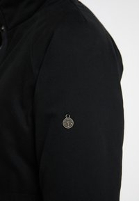 Sort stof overtøj med en tekstureret overflade, prydet med et rundt metalemblem på venstre ærme nær skulderen.