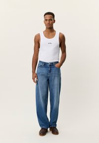 Άσπρο φανελάκι με μαύρο λογότυπο, συνδυασμένο με άνετο μπλε τζιν παντελόνι. Το μοντέλο στέκεται με τα χέρια στο πλάι, φορά καφέ παπούτσια.