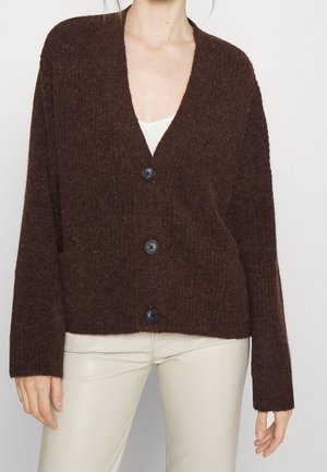 Bruine, textuurachtige cardigan met een diepe V-hals, lange mouwen en vier zwarte knopen aan de voorkant. Draagt over een lichte top en crèmekleurige broek.