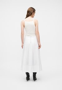 Γυναίκα με κασταν ά μαλλιά, που φοράει λευκό αμάνικο τοπ, μακριά λευκή φούστα με φερμουάρ στην πλάτη και μαύρα μπότες με τακούνι, στέκεται γυρισμένη προς τα πίσω.