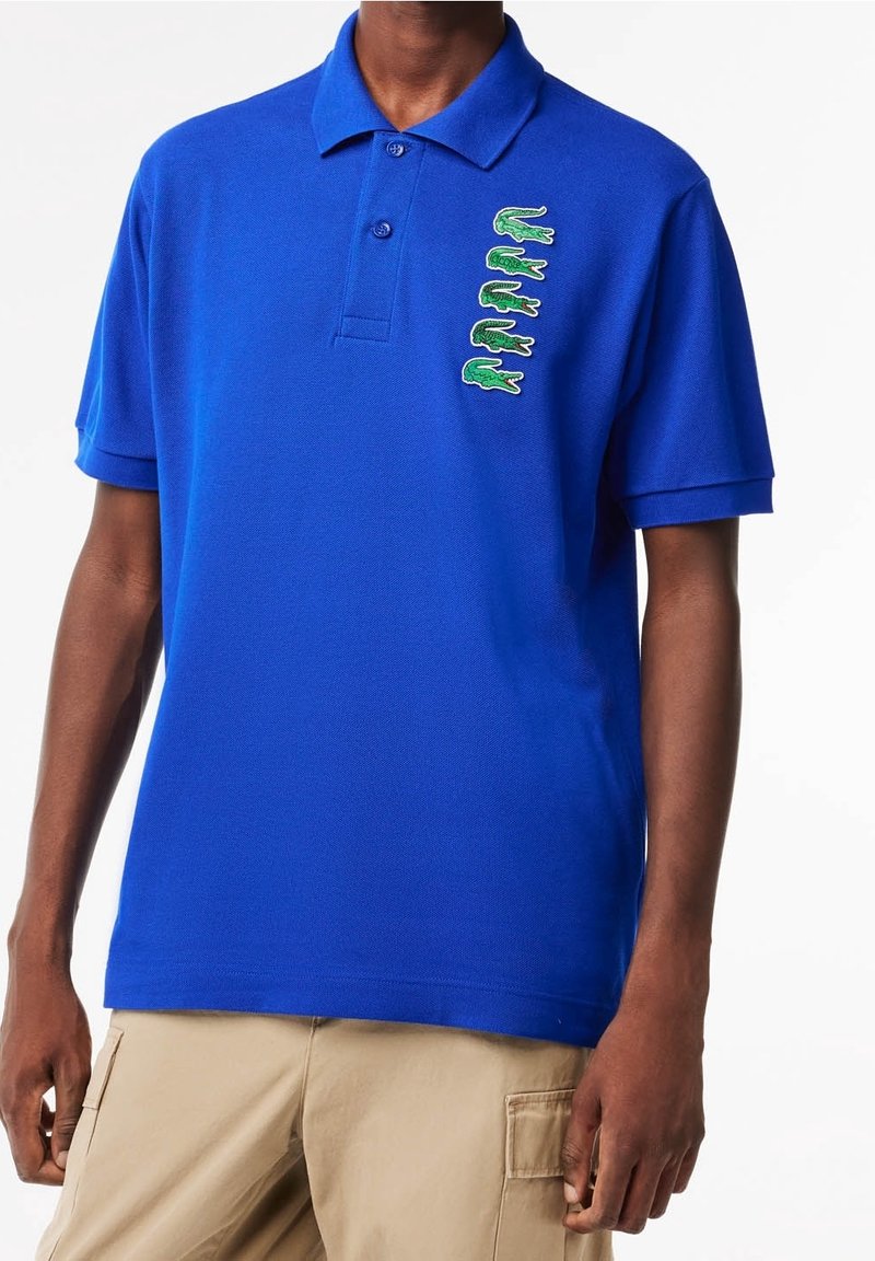 Polo bleu avec patte à deux boutons, manches courtes et broderie de crocodile verte en motif vertical sur le côté gauche de la poitrine.