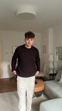 Pull en tricot noir avec motif texturé porté sur un pantalon ample de couleur crème. Intérieur aux tons neutres avec mobilier moderne.