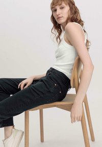 Weißes geripptes Tanktop, dunkelgrüne hochgeschnittene Jeans, Holzstuhl. Merkmale sind umgekrempelte Ärmel, seitliche Taschen und leichte Nähte.