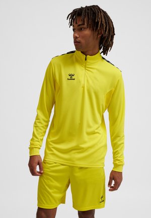 Homme portant une tenue de sport jaune vif avec un haut zippé à manches longues et un short, regardant sur le côté sur un fond uni.