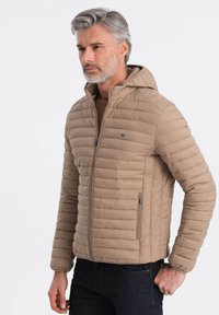 Beige pufferjakke med en lynlåshætte, der har et horisontalt quiltet design, to sidelommer og et lille logo på brystet.