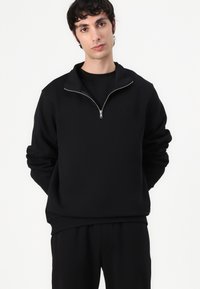Sudadera negra de pullover con cuello de media cremallera, textura suave, mangas largas y puños acanalados, combinada con pantalones negros.
