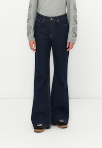 Jeans de ganga escura com corte flared, cinco bolsos, detalhamentos de costura sutis e design de cintura alta, conjugados com uma blusa de manga longa cinza.