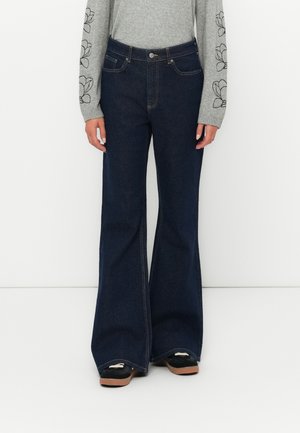 Jeans din denim albastru închis, cu fason evazat, cinci buzunare, detalii de cusătură subtile și un design cu talie înaltă, asortați cu un top gri cu mânecă lungă.
