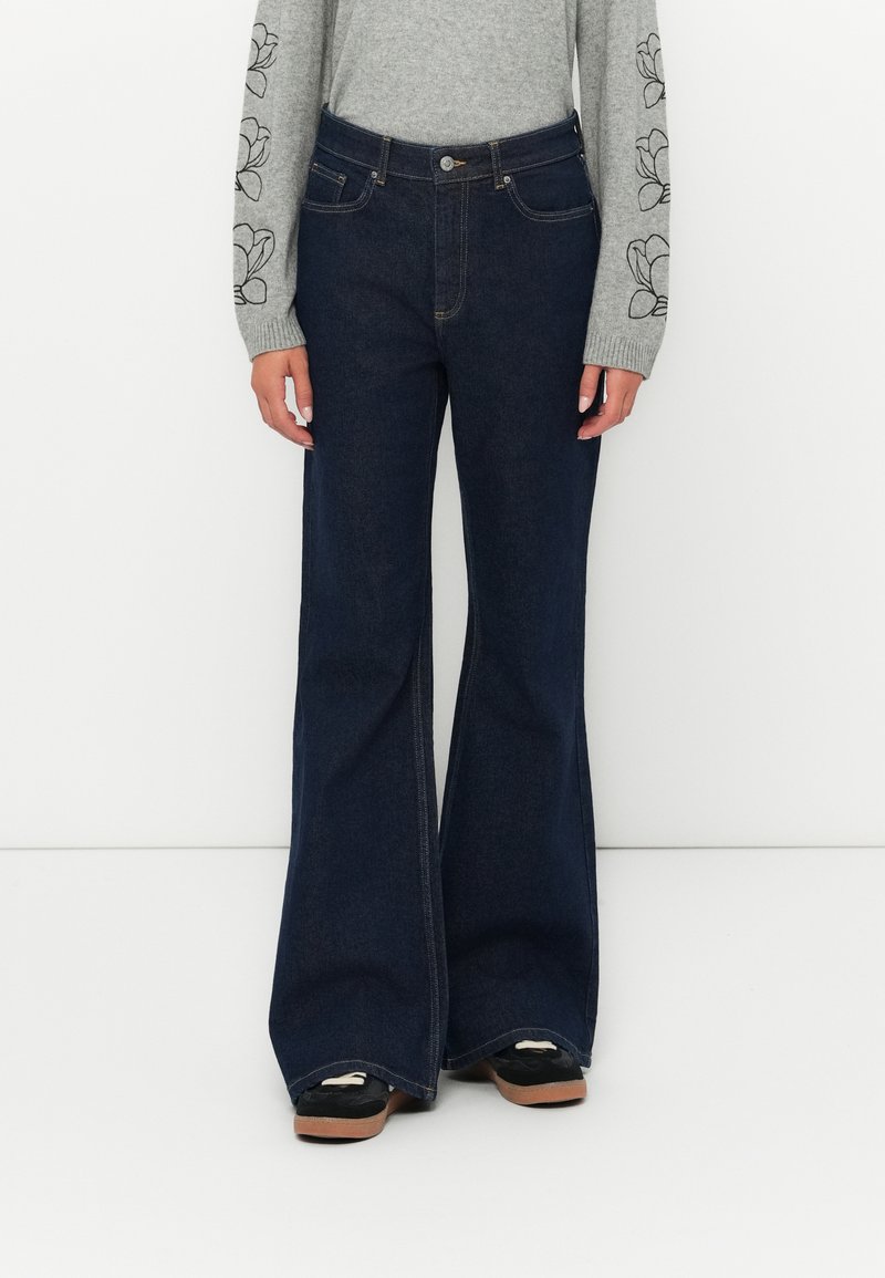 Jeans de ganga escura com corte flared, cinco bolsos, detalhamentos de costura sutis e design de cintura alta, conjugados com uma blusa de manga longa cinza.
