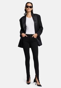 Czarna satynowa marynarka na białym t-shircie, zestawiona z czarnymi obcisłymi dżinsami z bocznymi zamkami i czarnymi wysokimi szpilkami.