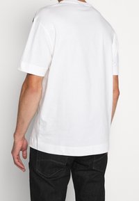 Bílý bavlněný tričko s krátkými rukávy, kulatým výstřihem a rovním lemem. Zadní strana je hladká bez viditelného loga nebo vzorů.