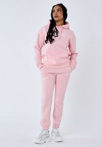 Sudadera rosa con bolsillo delantero y cordones, combinada con joggers rosas. Presenta puños de canalé y un logo bordado. Textura suave.