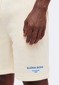 Σκούρο δέρμα χεριού που ακουμπάει σε κρεμολευκά σορτς με μπλε κείμενο "BJÖRN BORG STOCKHOLM 1956" κοντά στο τελείωμα.