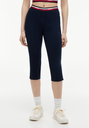 Kobieta ubrana w granatowe legginsy capri z czerwoną i białą gumą Tommy Jeans, białe skarpetki za kostkę i beżowe trampki, stojąca na białym tle.