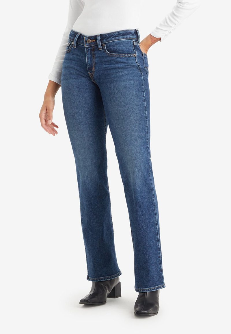 Donna che indossa jeans blu a vita media e gamba dritta, con una maglietta bianca a maniche lunghe e stivaletti neri con tacco, con una mano in tasca.