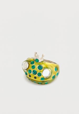 Saplu gredzens, kas veidots kā ķirzaka, spilgti dzeltenā krāsā ar zaļiem rhinestones un sudraba akcentiem, ar mazu kroņu augšpusē un lielām apaļām acīm.
