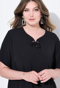 Top nero con scollatura a V e dettaglio annodato, maniche corte e texture liscia. La modella indossa orecchini d'argento texture di grandezza larga.
