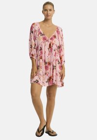 Una mujer se encuentra de pie con un vestido floral rosa y chanclas negras, mostrando un estilo de verano relajado y casual. Se presentan colores brillantes y alegres.