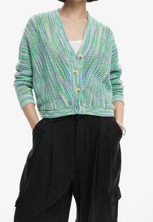 Person iført en pastelgrøn, lilla og blå strikket cardigan med knapper over en hvid top, kombineret med løse sorte bukser og et armbånd.