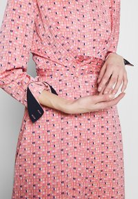 Femme portant une robe rose à motifs avec des poignets noirs, tenant une main délicatement sur l'autre à la taille.