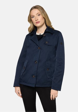 Kobieta w granatowej, zapinanej na guziki kurtce z kołnierzem, kieszeniami z patkami i długimi rękawami, stojąca na tle jasnego, jednolitego tła.