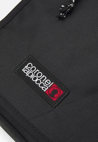 Bolsa de tela negra texturizada con un parche de logo negro y rojo. Cuenta con un cierre de cordón con un cordón negro trenzado.