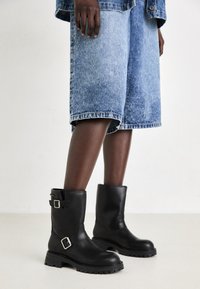 Botas de tornozelo em couro preto com sola grossa, apresentando fivelas prateadas e uma textura suave. Usadas com calções de ganga azuis.