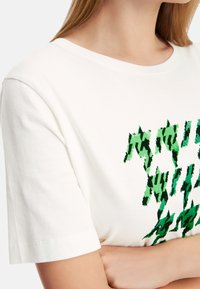 Weißes Baumwoll-T-Shirt mit kurzen Ärmeln, das ein grünes abstraktes Grafikdesign zeigt. Der Stoff hat eine glatte Textur und einen lässigen Schnitt.