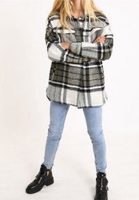 Giacca a camicia a quadri nei toni grigio, bianco e verde oliva, con chiusura a bottoni, due tasche sul petto e una vestibilità comoda. Abbinata a jeans blu e stivali neri.