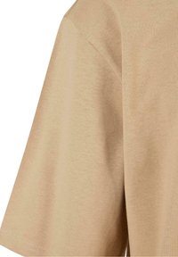 T-shirt beige en coton à manches courtes montrant la partie supérieure du bras et l'épaule avec des détails de couture visibles.