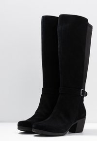 Stivali neri in suede al ginocchio con tacco a blocco, dotati di un dettaglio con cinturino e fibbia attorno alla caviglia. Superficie liscia, design con punta affusolata.