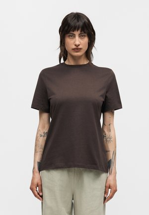 Femme aux cheveux mi-longs foncés, portant un t-shirt marron foncé uni à manches courtes et un pantalon beige clair, debout sur un fond gris.
