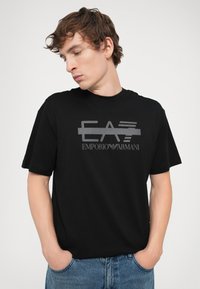 Black cotton t-shirt with short sleeves featuring a gray logo text "EA7 EMPORIO ARMANI" on the front, worn with blue jeans.
