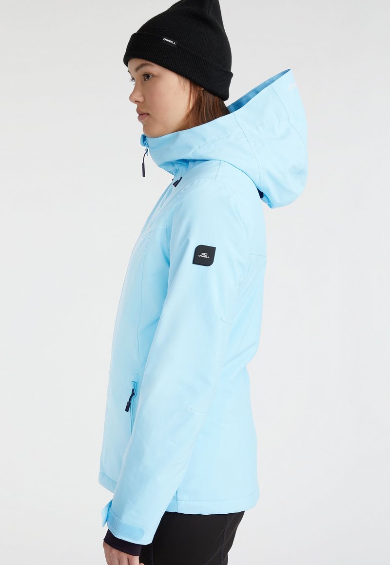 Giovane che indossa una giacca a cappuccio blu chiaro e un berretto nero, in piedi di lato su uno sfondo bianco semplice.