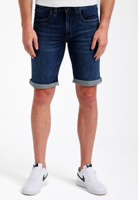 Pantalones cortos de mezclilla en azul oscuro con dobladillo enrollado, diseño ajustado y un ligero desvanecimiento. Combinados con zapatillas Nike blancas con detalles en negro.