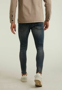 Jean bleu ajusté avec des zones décolorées et un tissu texturé, associé à des baskets blanches. La chemise est beige avec des détails de boutons sur les manches.