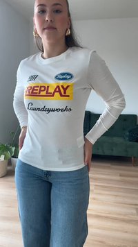 Camisa de manga longa branca com o logótipo "REPLAY" em vermelho e amarelo, texto preto, decote redondo, combinada com jeans de tom azul claro. Estilo casual moderno.