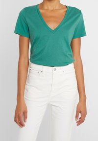 Donna che indossa una maglietta azzurro verdeflex con scollo a V e maniche corte, infilata in pantaloni bianchi a vita alta con chiusura a bottoni e zip.