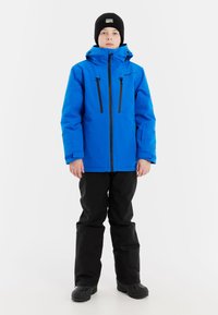 Blaue wasserdichte Jacke mit schwarzen Reißverschlüssen und Taschen, kombiniert mit schwarzen Hosen und Stiefeln. Ausgestattet mit einer Kapuze und einer strukturierten Strickmütze.