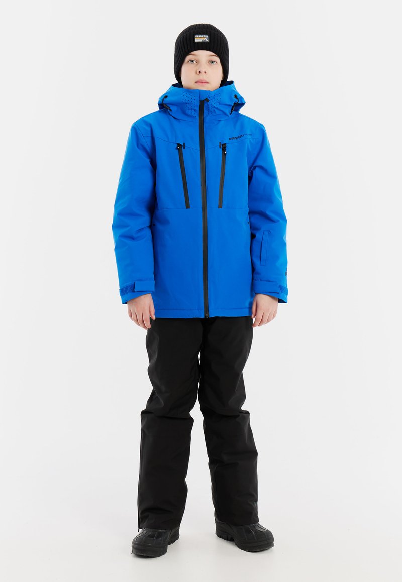 Blaue wasserdichte Jacke mit schwarzen Reißverschlüssen und Taschen, kombiniert mit schwarzen Hosen und Stiefeln. Ausgestattet mit einer Kapuze und einer strukturierten Strickmütze.