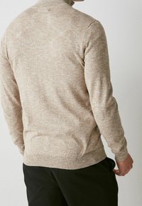 Beżowy sweter dzianinowy z wysokim kołnierzem, długimi rękawami i ściągaczami na mankietach. Tekstura wydaje się miękka i lekko melanżowa.