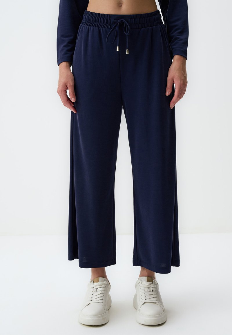 Marineblaue weit geschnittene Hose aus einem glatten, leichten Stoff mit einem elastischen Bund und Kordelzug. Kombiniert mit weißen Sneakers.