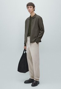 Giacca verde oliva con zip sopra una maglia a maglia verde scuro, abbinata a pantaloni crema a gamba larga e scarpe di pelle nere, con una borsa a tracolla nera.