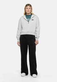 Nuori nainen seisoo kasvot eteenpäin, päällään vaaleanharmaa puoleen vetoketjuun avoin huppari, turkoosi graafinen t-paita, mustat housut ja valkoiset tennarit valkoisella taustalla.