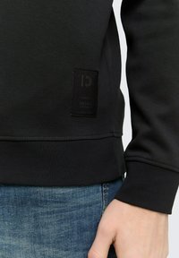 Sweatshirt negru texturat cu un detaliu subtil sub formă de patch logo, manșete ribbate și un design simplu, asortat cu blugi din denim spălati deschis.