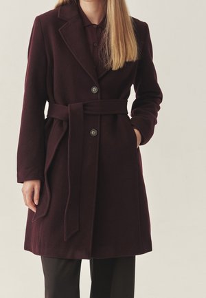 Vrouw draagt een donkergroene bordeauxrode wollen jas met ceintuur en knopen over een zwarte broek, met één hand in de zak, staand tegen een effen achtergrond.