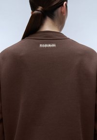 Brun sweatshirt med en slät textur. Har en logotypetikett på övre delen av ryggen. Enkel, avslappnad design utan ytterligare detaljer.